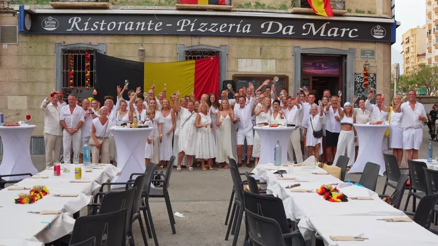 Vista elevada de la calle del restaurante: el rótulo «Ristorante - Pizzería Da Marc» en la fachada, una gran bandera de Bélgica colgada en la pared junto a la puerta, una bandera de España en un balcón y, delante, un grupo numeroso de personas vestidas de blanco brindando entre mesas altas con mantel.