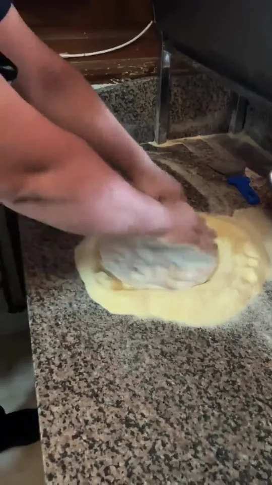 Dos manos aplastan y estiran un disco de masa sobre nuestra encimera de granito y le dejan el borde alto. Al lado, el bol de salsa de tomate y una rasqueta azul.