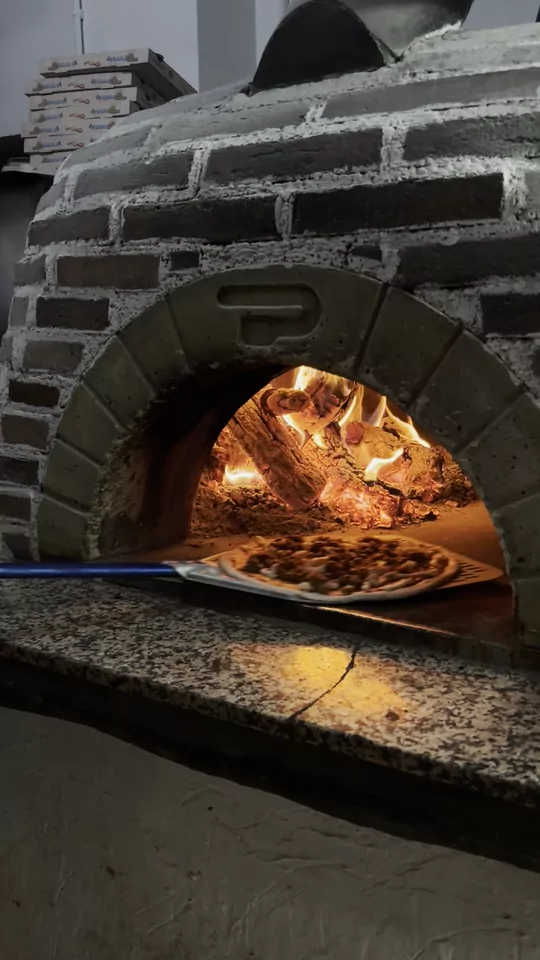 Una pizza cruda sobre la pala de mango azul entra en nuestro horno de leña: el arco de ladrillo con una marca en relieve en la clave, dentro el fuego naranja ardiendo sobre la solera, y el reflejo de la llama en la encimera de granito.