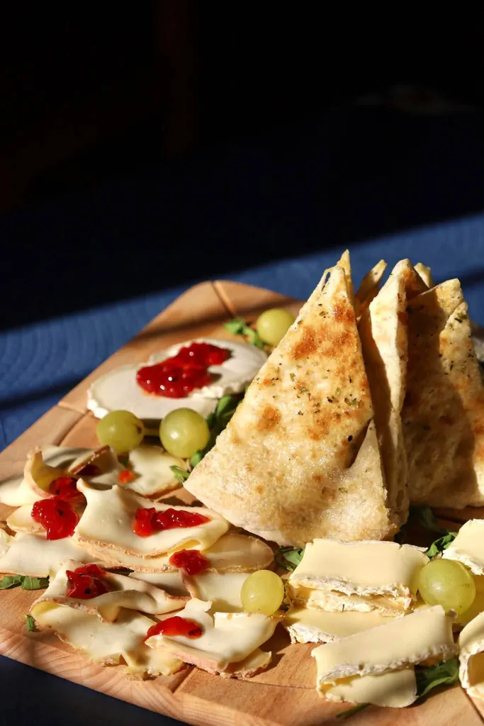 Una tabla de madera sobre mantel azul oscuro con triángulos de focaccia dorada puestos de pie, lonchas de queso y de fiambre extendidas alrededor, uvas verdes, hojas de rúcula y un queso redondo cubierto de mermelada roja.