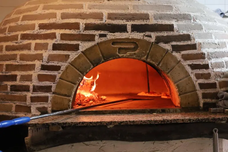 Nuestro horno de leña encendido: cúpula de ladrillo visto, encalada por el flanco derecho; arco de dovelas con una marca en relieve en la clave y, dentro, el fuego naranja sobre un lecho de brasa y ceniza; una pala de mango azul entra por la boca.