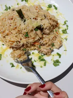 Plato blanco con arroz de grano largo en salsa cremosa, con trozos de pollo, tiras de zanahoria, verdura verde y queso rallado por encima. Alrededor, perejil picado y gotas de aceite.