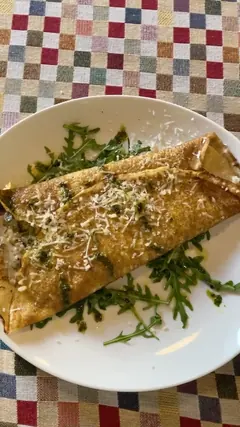 Crep doblado en dos, dorado y con burbujas tostadas, sobre un plato blanco: lleva queso rallado por encima, un hilo de aceite verde con hierbas y va apoyado en un lecho de rúcula. Por el borde izquierdo asoma un relleno cremoso con trozos oscuros. El plato está sobre el mantel de cuadros de colores.