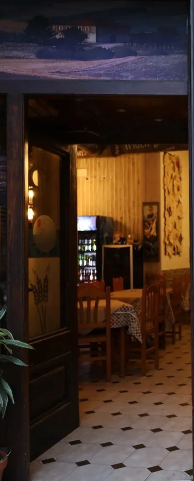 De noche, desde la acera: la puerta de madera oscura abierta y, por el hueco, el comedor iluminado en tonos cálidos —sillas de madera, mesas con mantel de cuadros, suelo de baldosa blanca y negra, una nevera de bebidas, un cuadro y un farol octogonal encendido—. Sobre el dintel, un mural pintado de un caserío entre campos. En primer plano, una planta en su maceta.