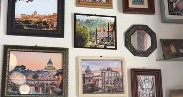 Una pared blanca llena de cuadros enmarcados con vistas italianas —un pueblo en una ladera, una casa de campo entre cipreses, una cúpula sobre un río al atardecer, unas ruinas y una galería cubierta de cristal— y, entre ellos, un espejo octogonal de marco tallado oscuro.