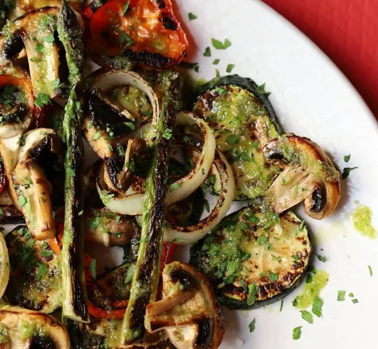 Plato blanco con verduras a la brasa, con sus marcas de parrilla: rodajas de calabacín, champiñones abiertos, espárragos verdes, tiras de pimiento rojo, aros de cebolla y berenjena, con un aliño verde de aceite y hierbas y perejil picado por encima.