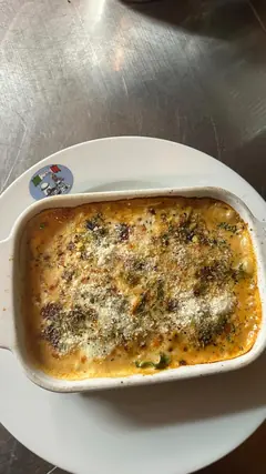 Fuente rectangular blanca con pasta gratinada al horno: la superficie dorada y burbujeante, queso rallado, orégano y trozos de verdura verde. Va sobre un plato blanco que lleva pegada la pegatina redonda del logotipo de la pizzería, en una encimera de acero.