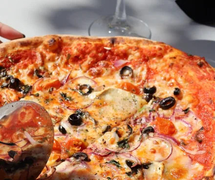 Una pizza recién hecha vista muy de cerca sobre la mesa: borde alto y tostado, tomate, queso fundido, aceitunas negras, aros de cebolla roja y trozos de tomate; por la esquina entra el cortador de rueda.