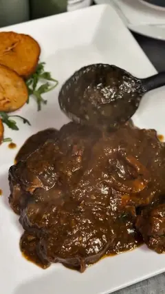 Plato cuadrado blanco con carne guisada en salsa oscura; un cazo negro deja caer más salsa por encima. A un lado, láminas de patata frita y una ramita de hoja verde.